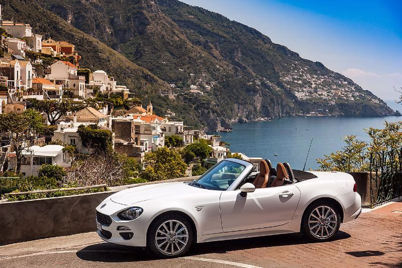 A white convertible parked above the Amalfi Coast with Positano and the cliffs descending towards the sea in the background.