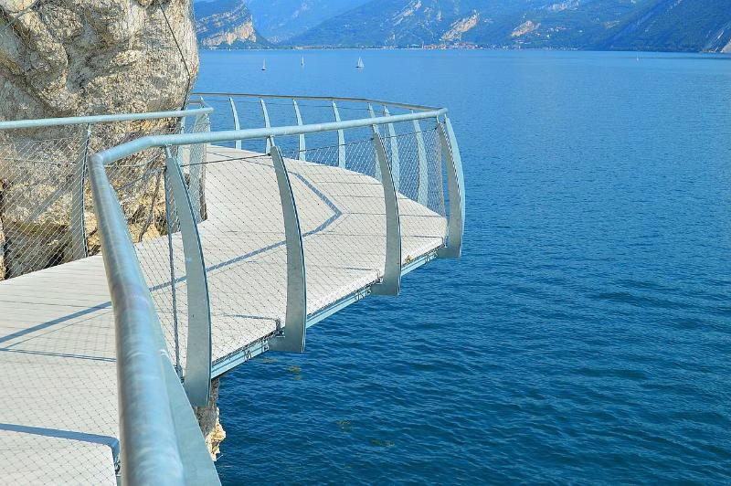 Suspended lakeside walkway of the Ciclabile di Limone extending over the blue waters of Lake Garda on a clear day.