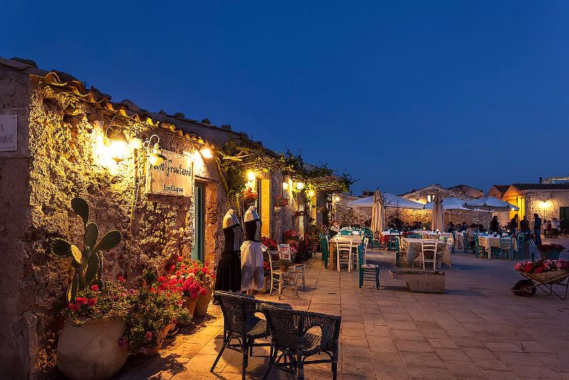 Evening scene in Marzamemi’s historic fishing square, with stone buildings, outdoor tables and warm lights at dusk.
