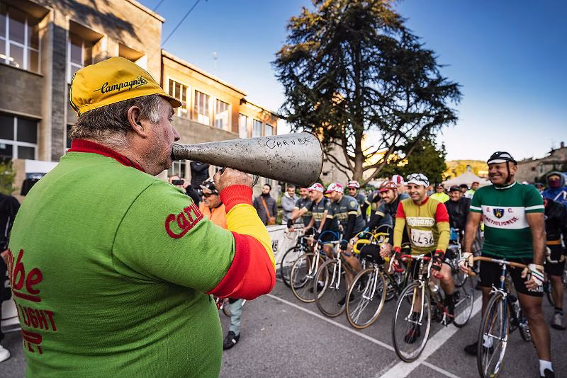 Starter calling riders to the beginning of L’Eroica vintage cycling event in Gaiole in Chianti, with participants in retro jerseys and classic steel bicycles.