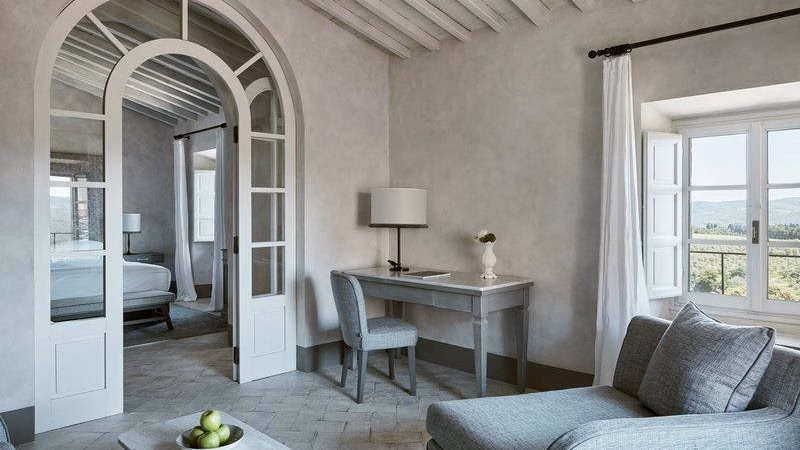 Tavarnelle Suite at Castello del Nero, featuring soft neutral tones, traditional beams, arched glass doors, and panoramic views over the historic Chianti estate.