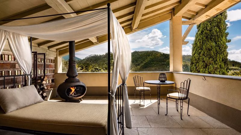 Deluxe Aurora Suite terrace at Locanda al Colle, featuring a canopy daybed, outdoor fireplace, and panoramic hillside views over the Versilia countryside.
