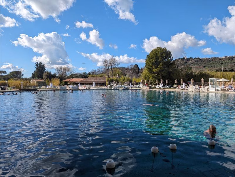 Guests bathing in the large thermal spring pool at Terme di Saturnia with hills and blue sky in the background