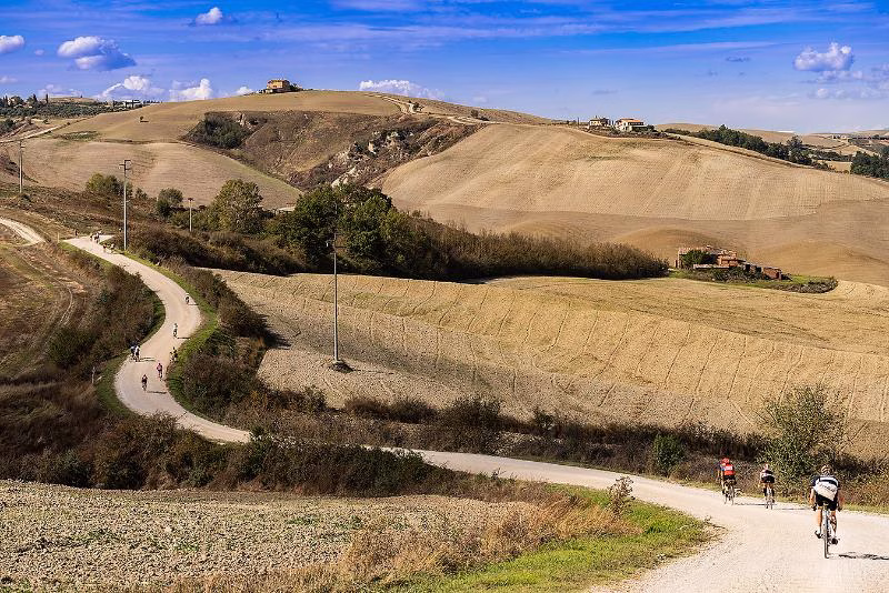 Cyclists riding along a winding white road through Chianti’s rolling hills and open farmland under a bright blue sky.
