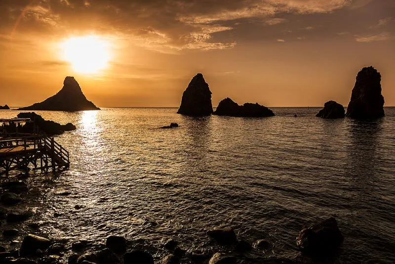 A golden sunset over the Faraglioni sea stacks of Aci Trezza, with calm waters reflecting the light and a wooden pier extending into the Ionian Sea.