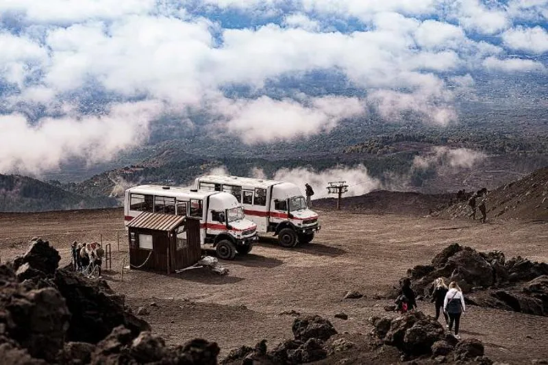 Off-road vehicles parked on the rugged volcanic terrain of Mount Etna, with hikers exploring the landscape and a sea of clouds rolling over the valley below.