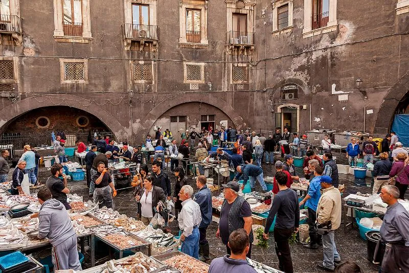 The bustling Pescheria fish market in Catania, with vendors selling fresh seafood, locals haggling, and historic lava-stone buildings in the background.