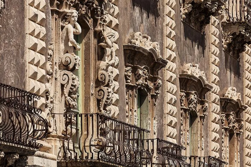 The intricately carved baroque façade of Palazzo Biscari in Catania, featuring ornate stone decorations, wrought-iron balconies, and detailed sculptural reliefs.