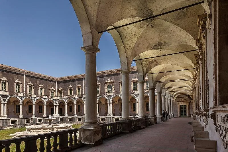 The grand cloister of the Monastero dei Benedettini in Catania, featuring elegant arched colonnades, a central courtyard with a fountain, and baroque architectural details.