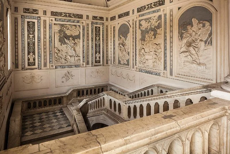 The grand staircase of Palazzo Biscari in Catania, adorned with intricate stucco reliefs, classical motifs, and elegant marble balustrades.