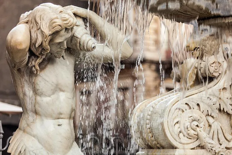 A marble statue of a youthful male figure atop the Fontana dell’Amenano in Catania, holding a large fish, with water cascading over the fountain’s edge.
