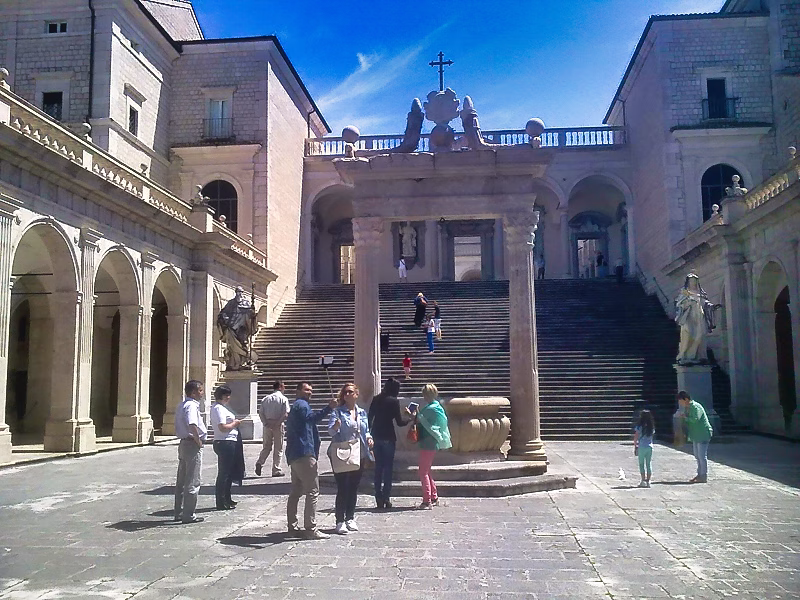 Visitors in the cloistered courtyard of Montecassino Abbey, gathered by the central well with the grand staircase and statues beyond.