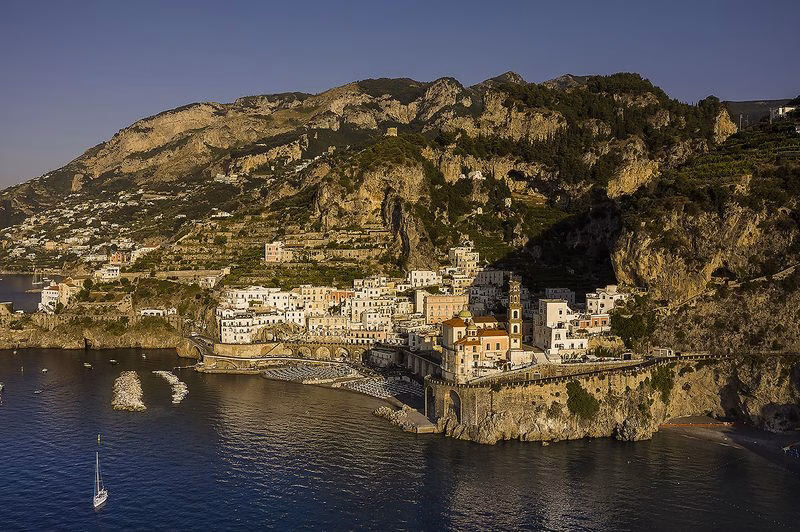 Wide view of Atrani’s cliffside village and beach with the Church of Santa Maria Maddalena, breakwaters and terraced mountains in late-afternoon light on the Amalfi Coast.