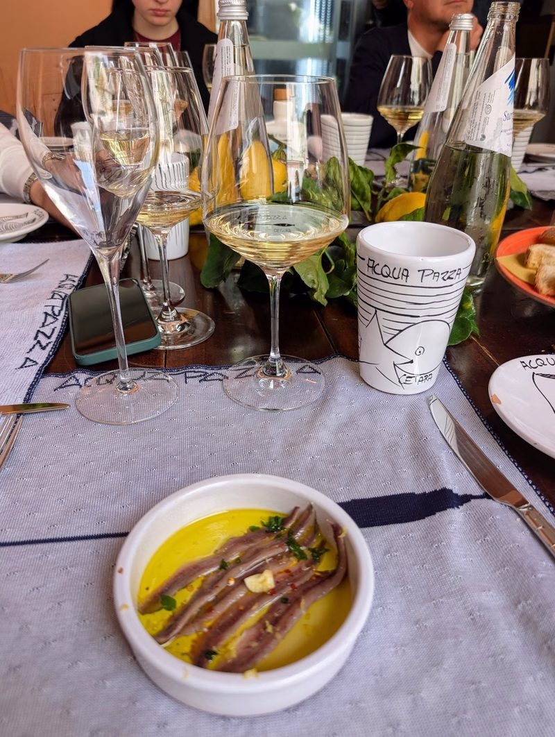 Small dish of anchovy fillets in extra-virgin olive oil with garlic and herbs on a restaurant table set with white wine glasses; “Acqua Pazza” cup visible.