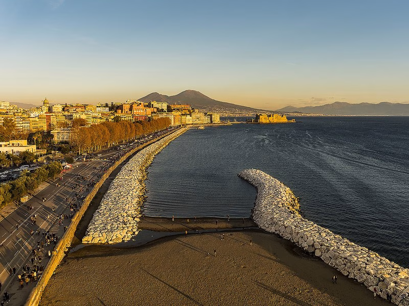 Aerial view of the Lungomare Caracciolo and Mergellina promenade with Castel dell’Ovo glowing at sunset and Mount Vesuvius on the horizon.