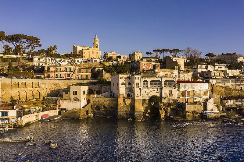 Terraced houses and arches of Marechiaro above the sea, with the bell-tower of Santa Maria del Faro and umbrella pines at golden hour.