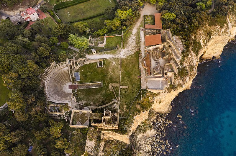 Aerial view of the Parco del Pausilypon in Posillipo, showing the Roman theatre, terraces and cliff edge above clear blue water; access via the Seiano Grotto tunnel.