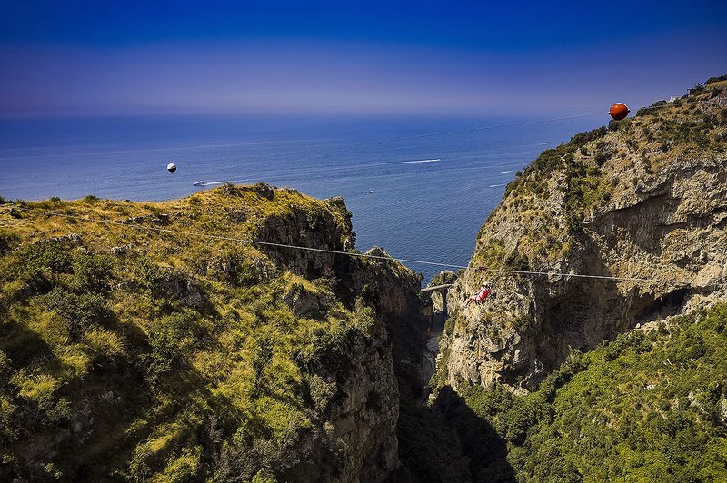 Rider on the Fiordo di Furore zipline crossing a rocky gorge high above the Amalfi Coast, with the bridge and open Tyrrhenian Sea in the background.