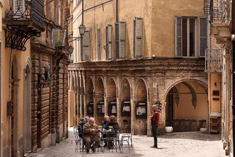 Café scene in Offida historic centre with brick arcades, shuttered façades and locals seated outdoors