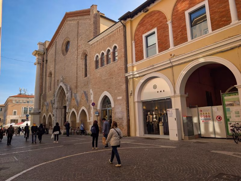 People walking past Pesaro Cathedral along a pedestrian street in the historic centre, with Romanesque brick architecture and arcaded buildings.