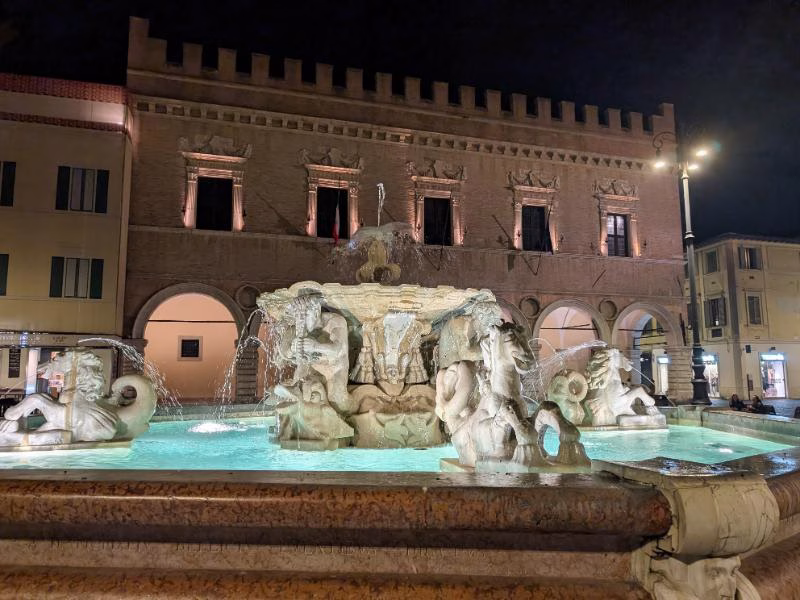 Illuminated fountain in Pesaro’s main square at night, with the Palazzo Ducale in the background and arcaded façades lit by warm evening lights.