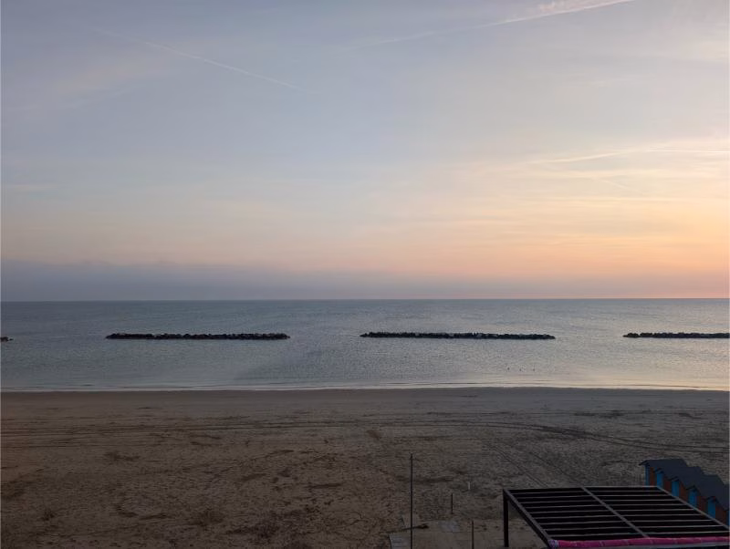 Wide sandy beach in Pesaro at sunset with a calm Adriatic Sea, pastel sky and offshore breakwaters on the horizon.