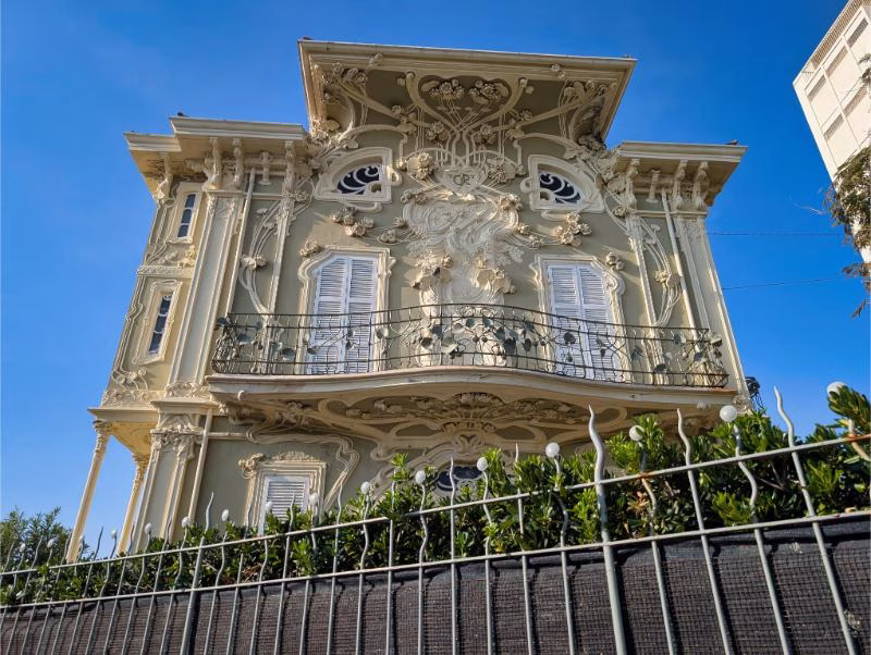 Ornate Liberty-style villa in Pesaro with sculpted floral decorations, curved iron balcony and pale green façade under a clear blue sky.
