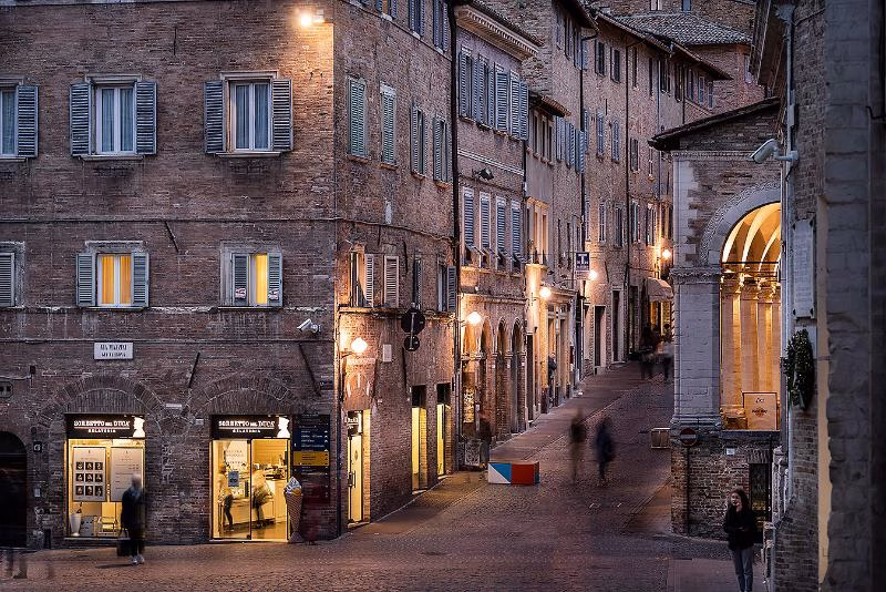 Evening street scene in Urbino historic centre with brick buildings, shuttered windows and softly lit shops