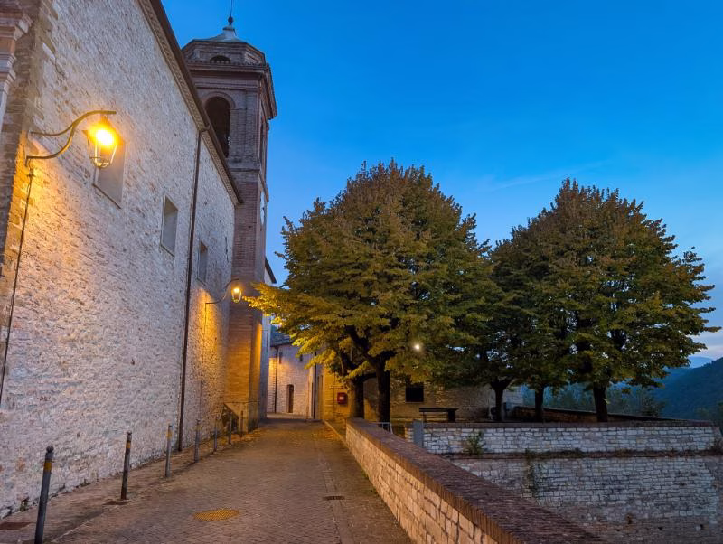Narrow lane beside a stone church in Genga, with trees and soft evening light overlooking the valley