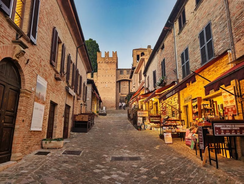 Evening view of Gradara’s main street leading up to the castle, lined with brick houses, shops and warm lights.