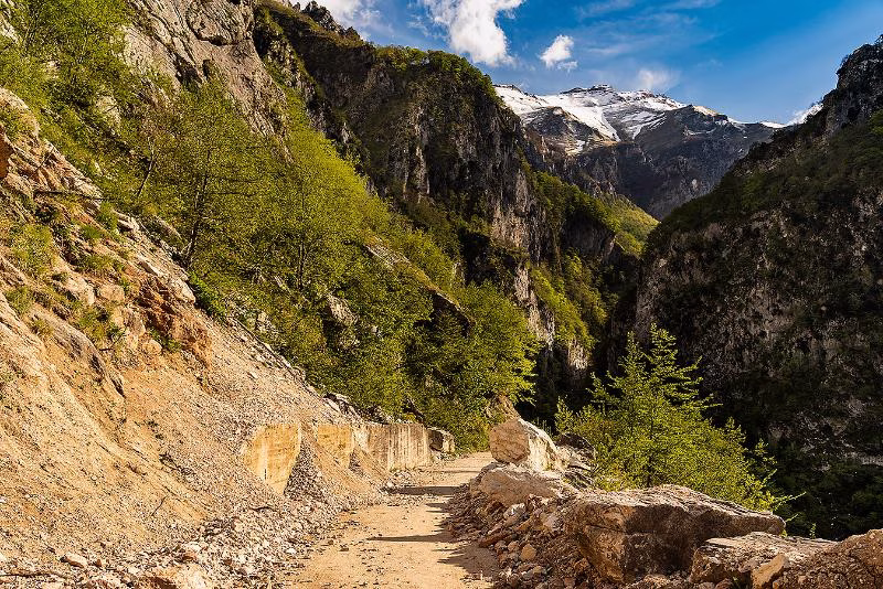 Mountain path cutting through the Frasassi Gorge in northern Le Marche, with rocky cliffs, spring greenery and snow-capped peaks in the distance.