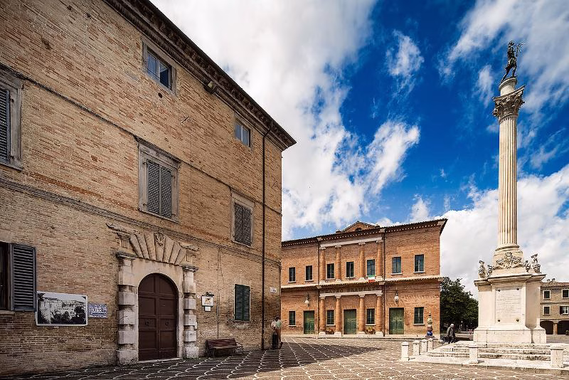 Brick piazza in Urbania, with theatre façade and tall column under a bright blue sky.
