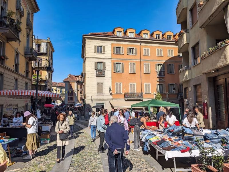 Busy street market in Turin’s Aurora district, with stalls, shoppers, and pastel apartment façades in bright sunlight.