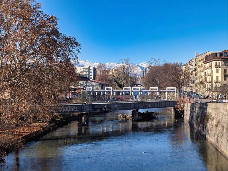 River Dora Riparia in Turin near Aurora, with a bridge, winter trees, and snow-capped Alps in the distance.