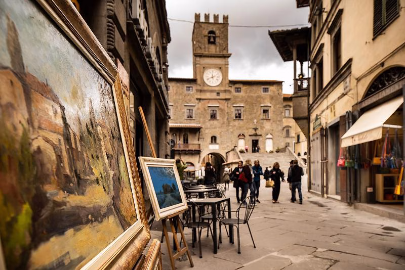 A gentler cultural tempo Street in a Tuscan town with easel paintings and a clock tower in the distance