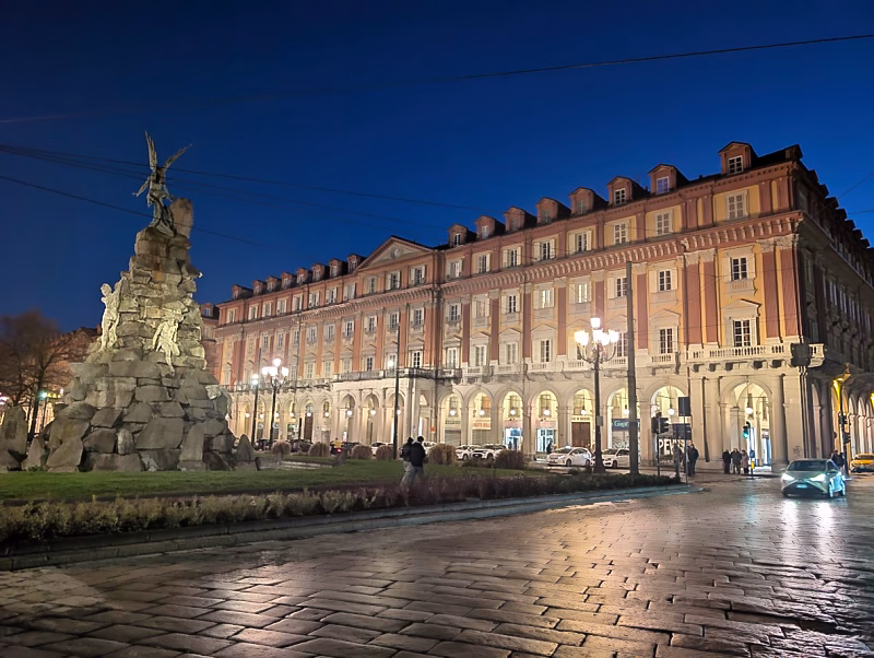 Piazza Statuto in Turin at night, with the Monumento al Traforo del Frejus and arcaded buildings.