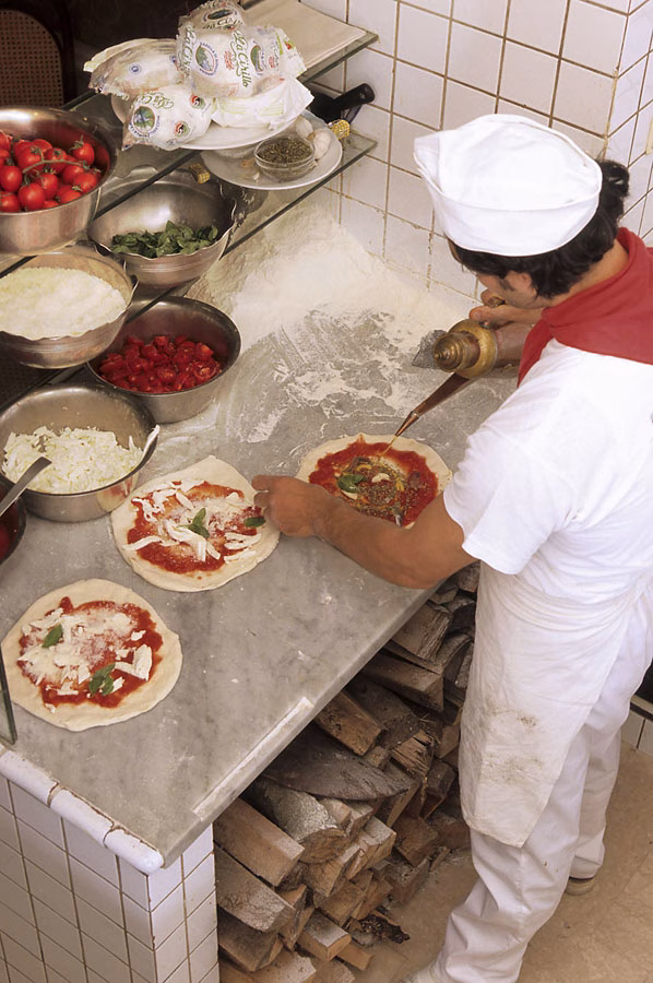 A pizzaiolo adding oil to pizzas beside bowls of tomato, basil and cheese