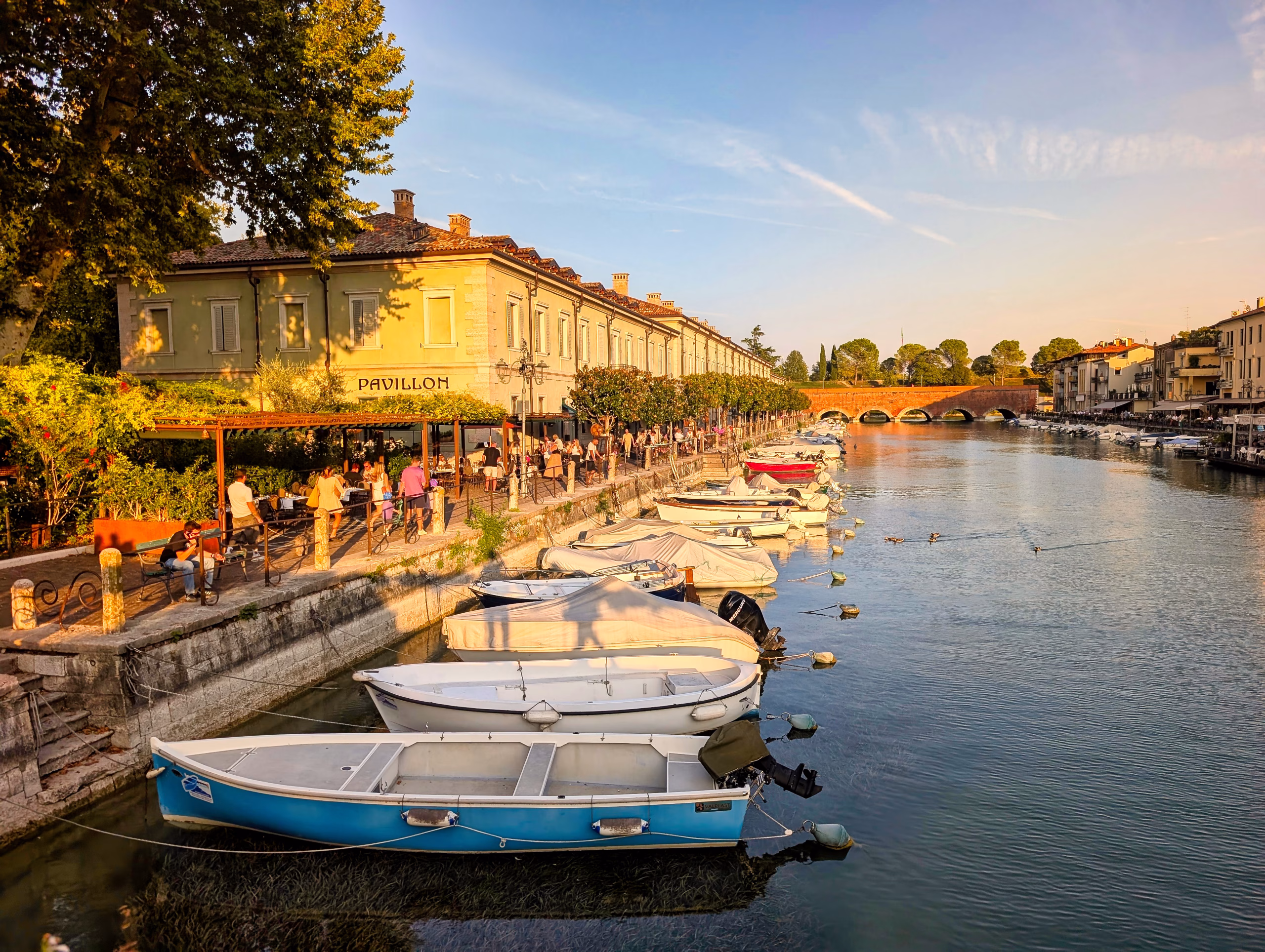 Small boats moored beside the tree-lined canal in Peschiera del Garda in late-afternoon light