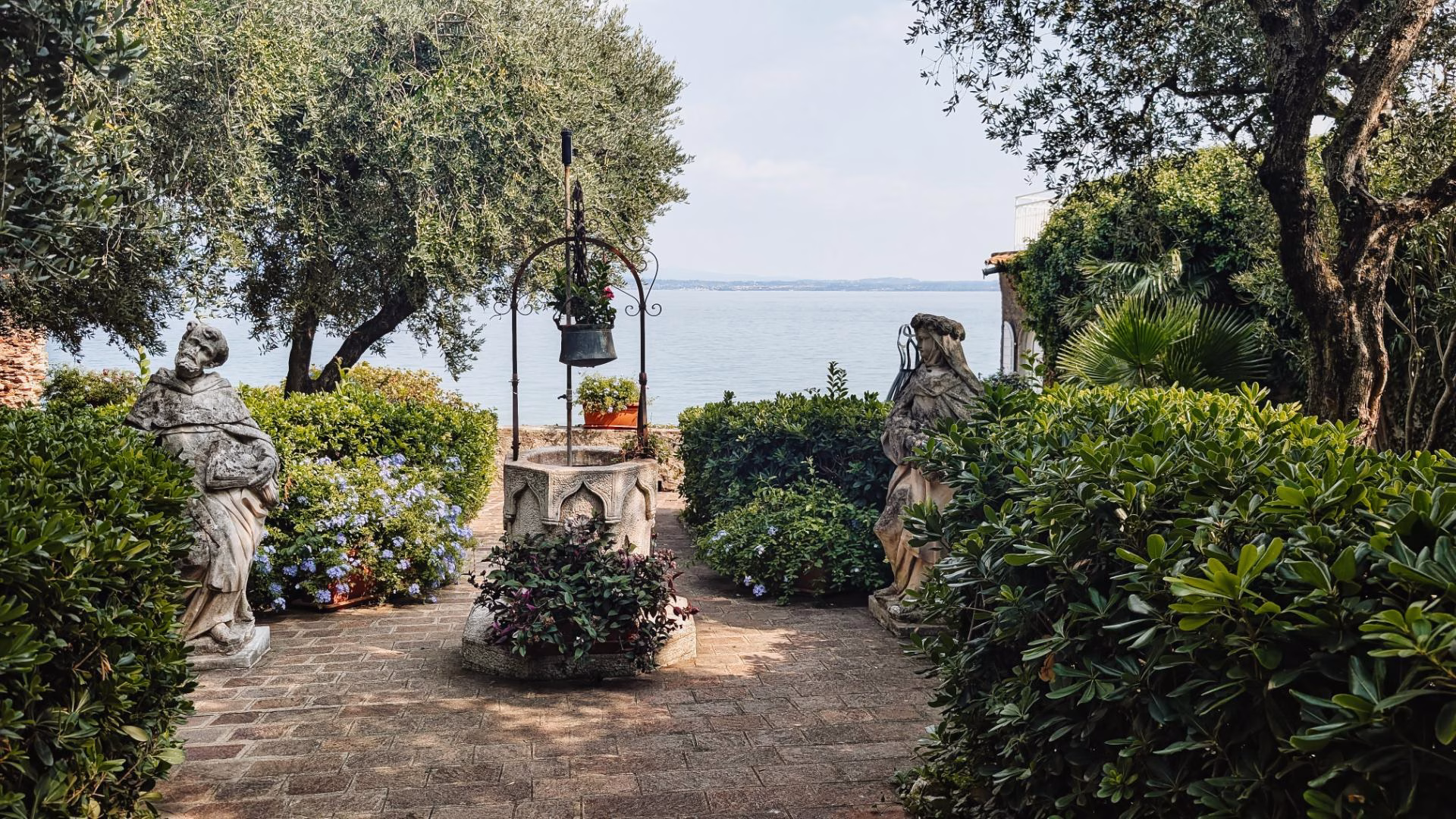 A lakeside garden, with olive trees, statues and a stone well.